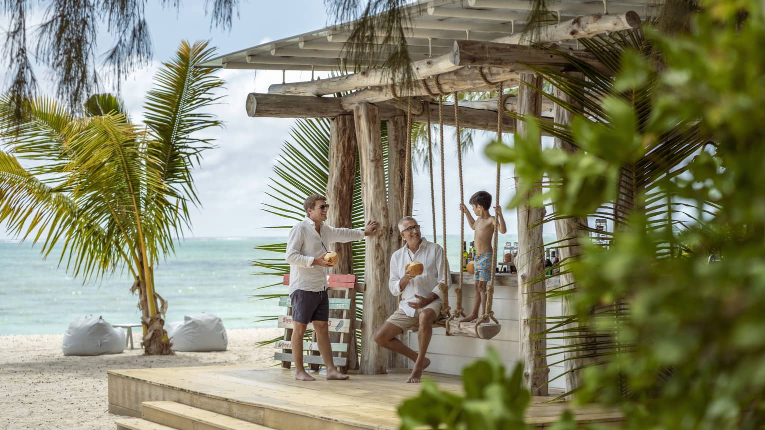 Two men smile at a boy standing on a beach swing at Ile Aux Cerfs