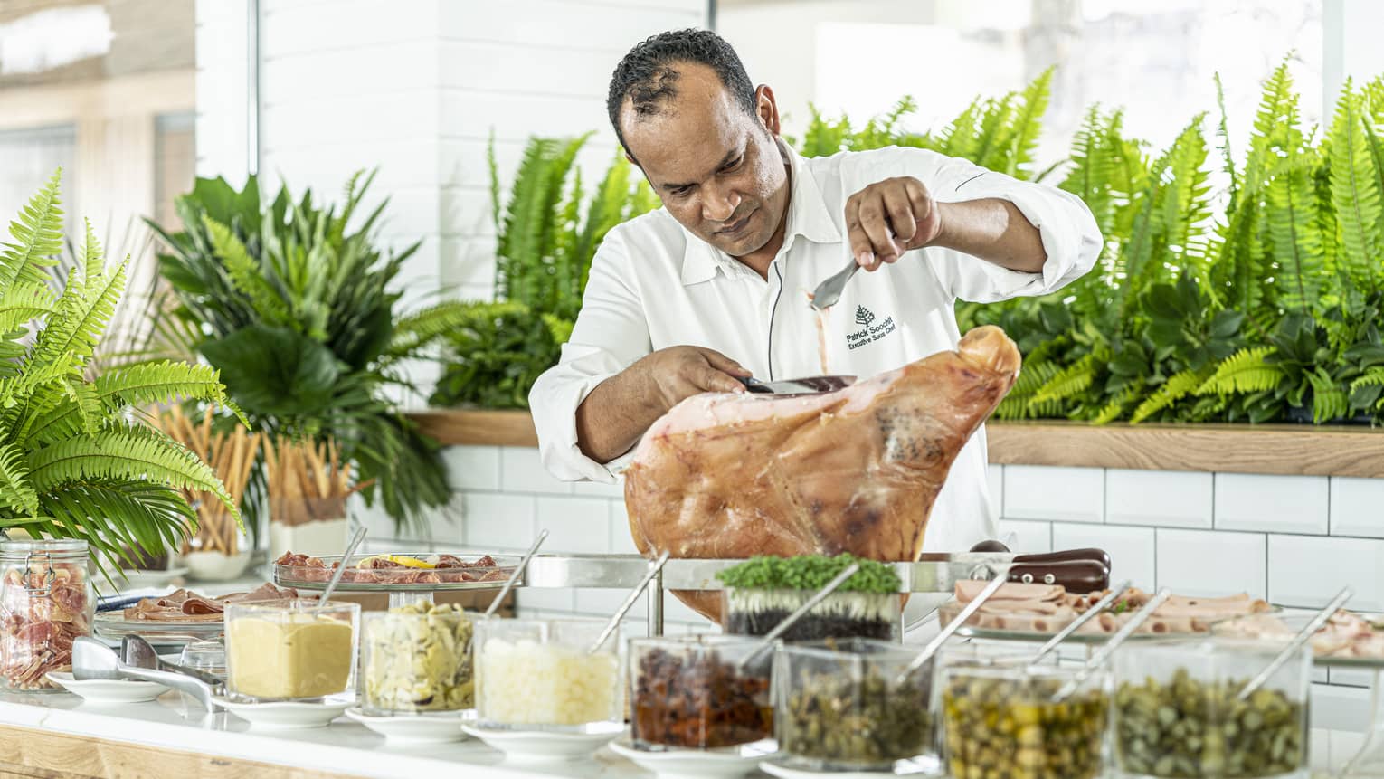 Chef in white shirt slicing meat on counter behind row of condiments in dishes