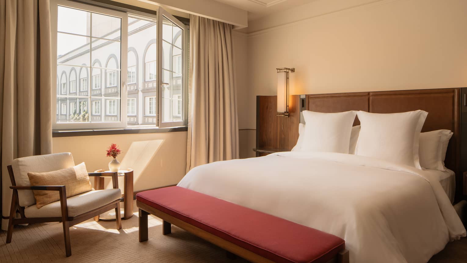 Hotel room with king bed, red bench, arm chair and window looking out to courtyard