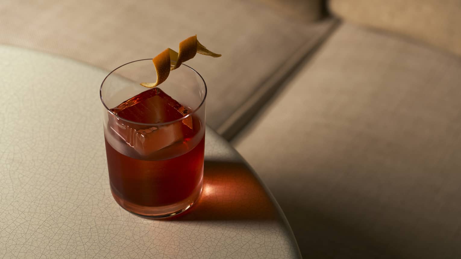 At a table's edge sits a negroni cocktail garnished with a twisted orange rind, light shining through its one large ice cube.