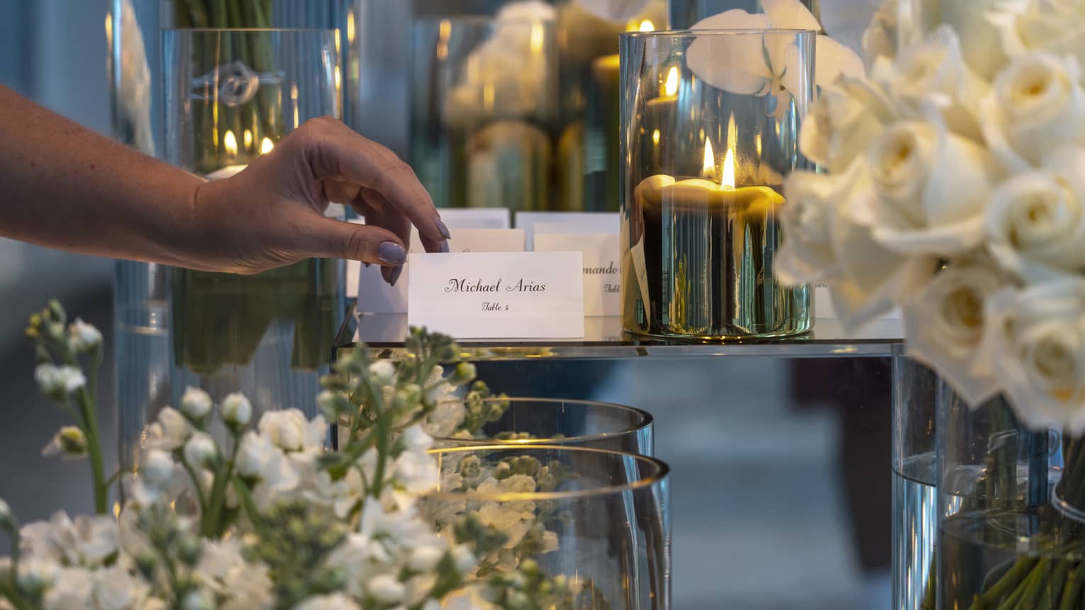 A close-up of a wedding guest picking up a place card from an elegant display at Four Seasons. The table features a sophisticated arrangement of white roses, with golden candle votives softly illuminating the background, creating a warm and welcoming ambiance for the celebration.