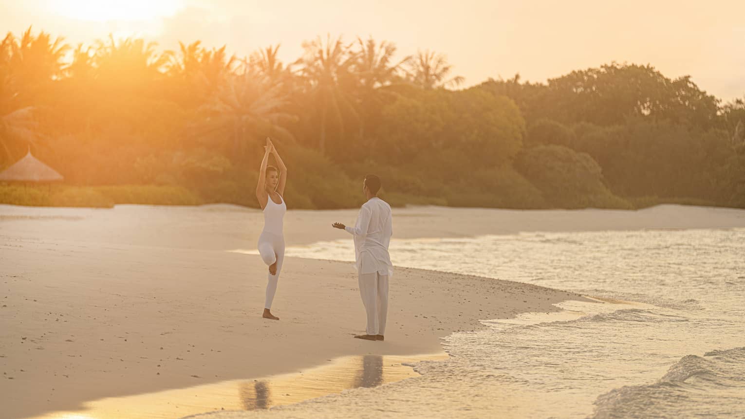 Person practicing yoga on a tropical beach at sunrise with an instructor, surrounded by calm ocean waves and lush greenery in the background