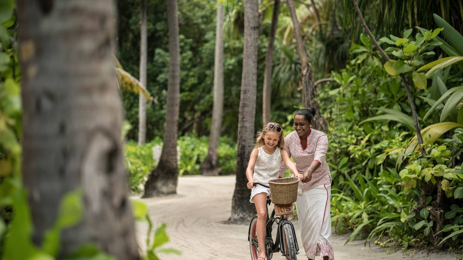 Hotel staff helps young girl ride bicycle with woven basket on tropical resort path