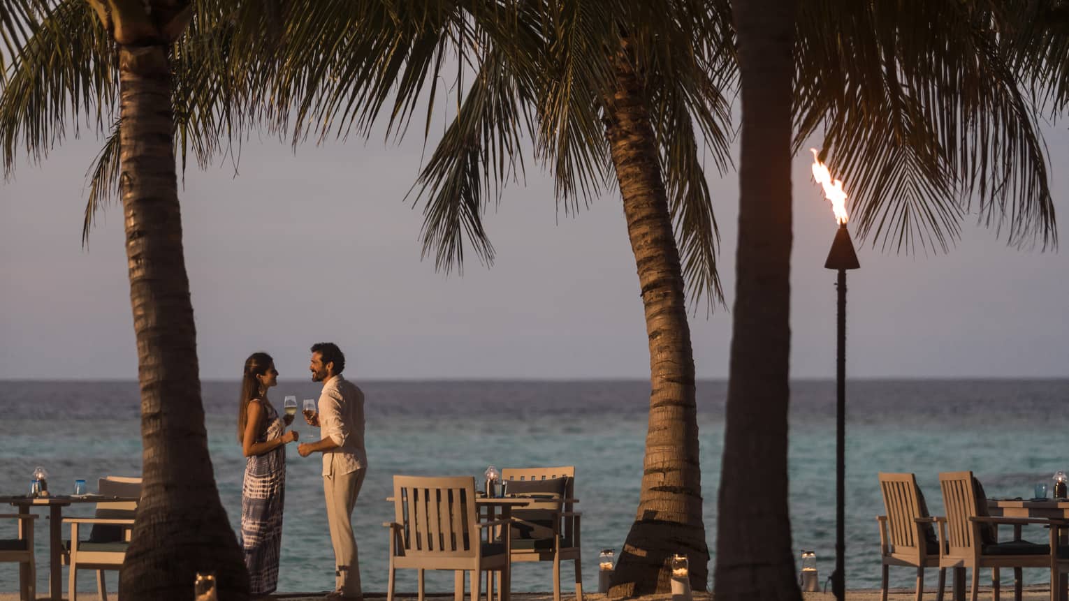 A couple talks on the beach at sunset, a tiki torch burns, table and chairs sit on beach