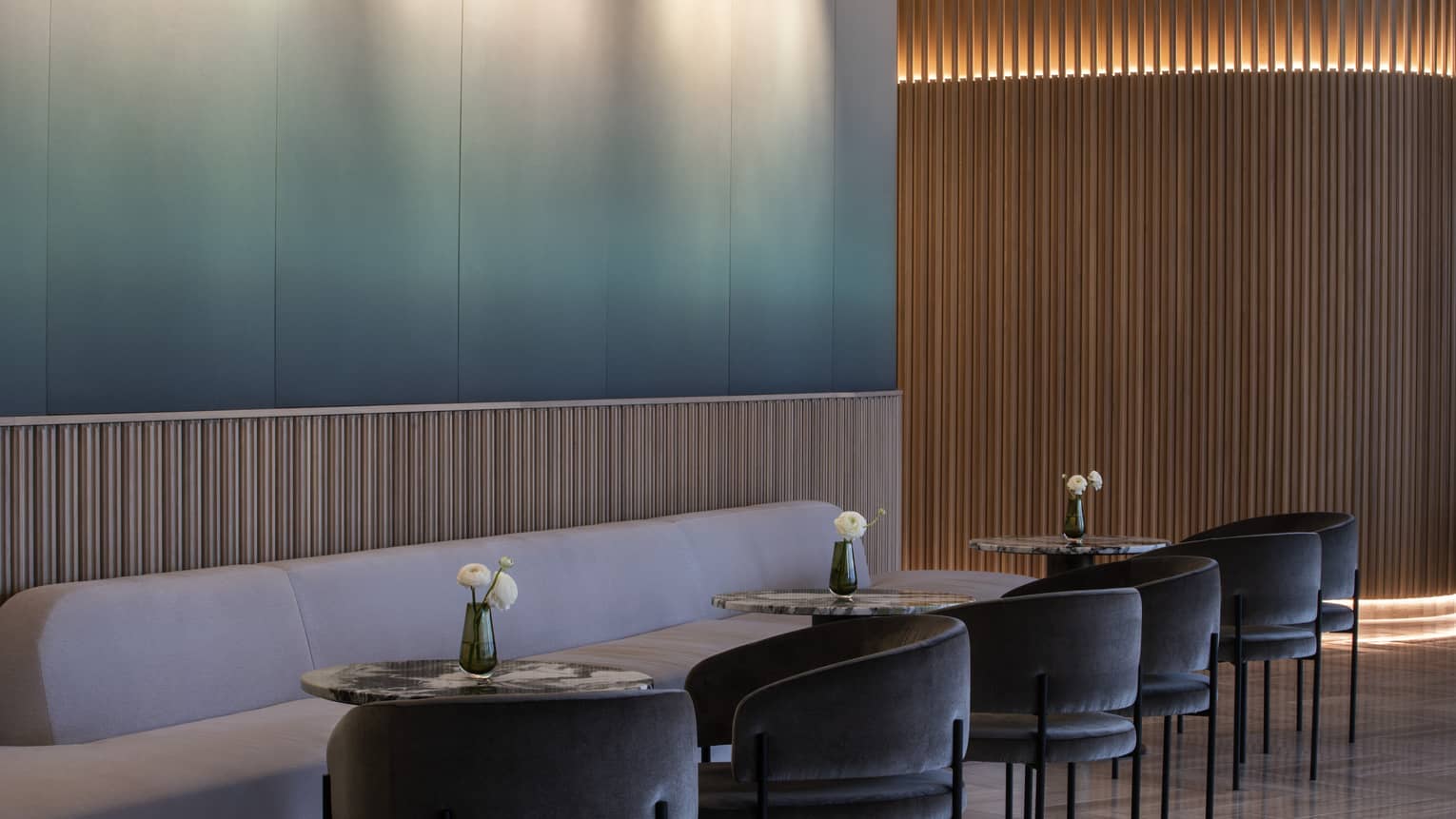 Blue-to-white gradient wall, light-grey plush banquet, three round cocktail tables and six dark-grey curved chairs at MARCUS Lounge
