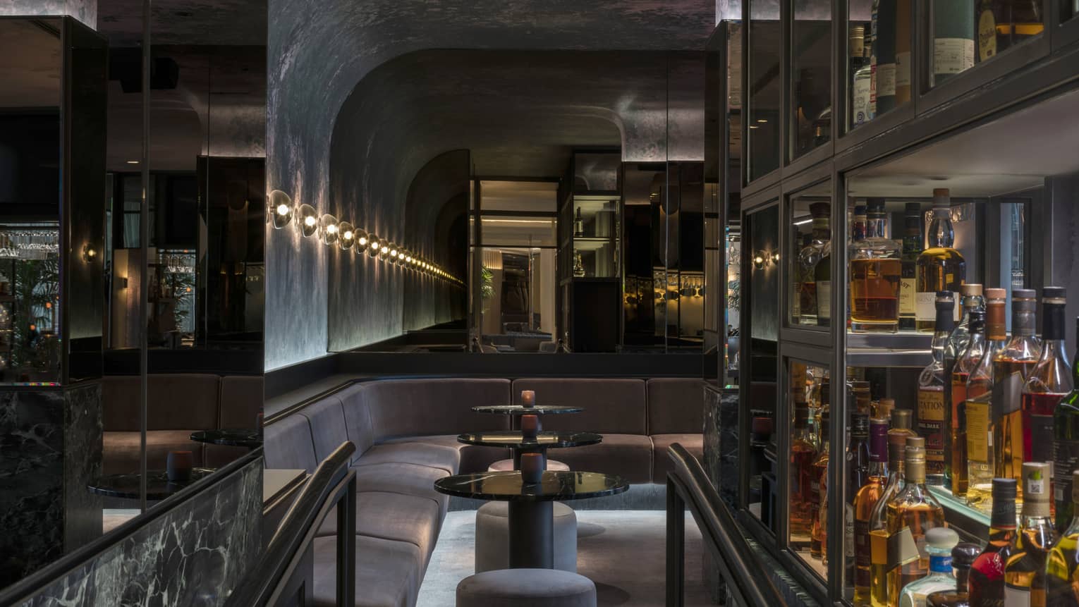 A velvet banquet and three round cocktail tables line the curved wall of MARCUS bar next to glass shelves holding bottle of spirits, all dimly lit