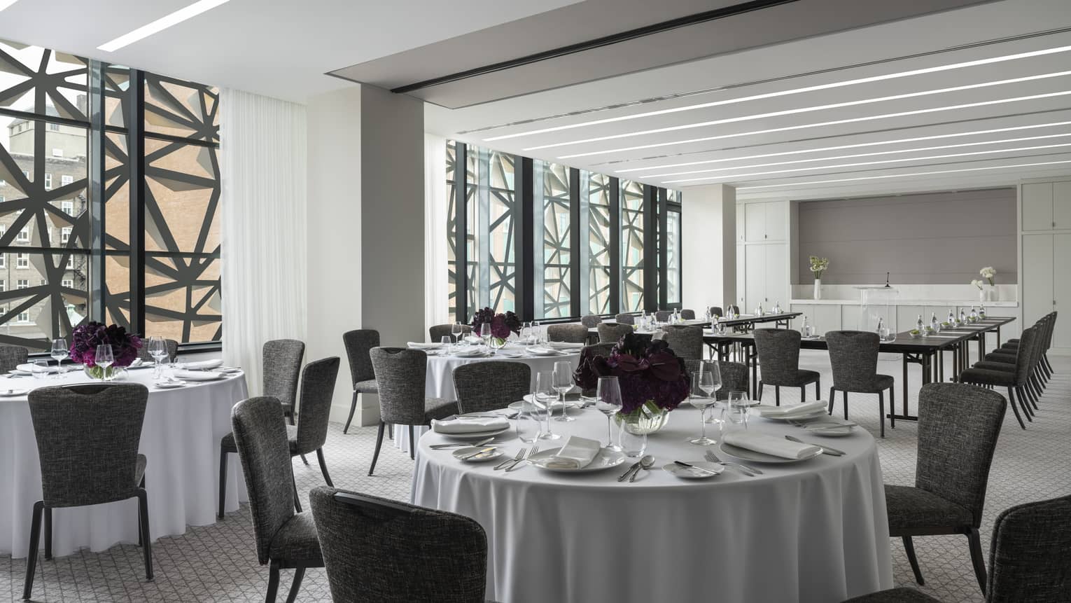 Salon with white round tables and flowers, grey chairs, U shaped table in background, ceiling-to-wall windows