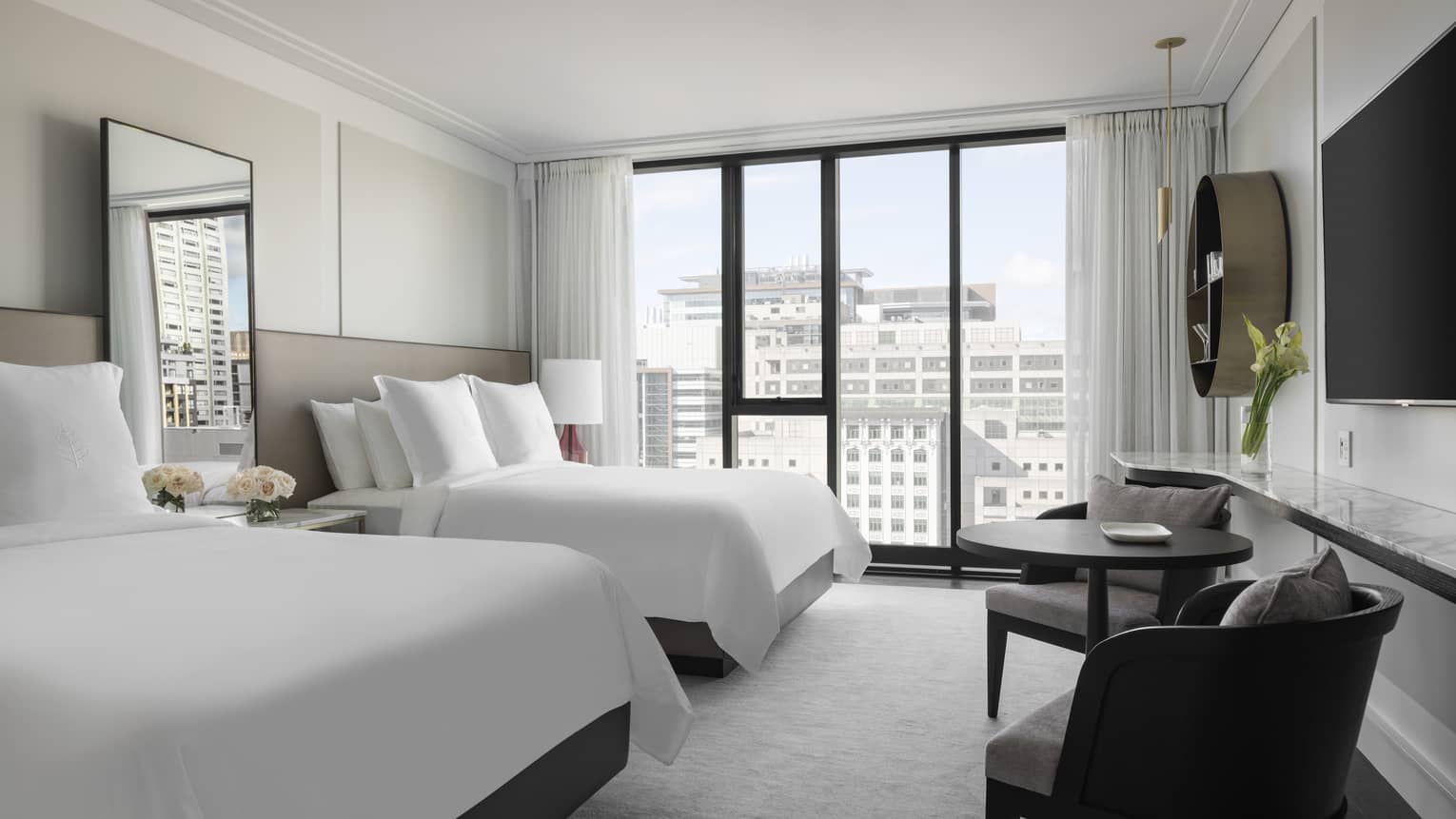 A bright hotel room with two double beds featuring white linens, large windows with city views, wall-mounted TV, and small seating area with table and chairs