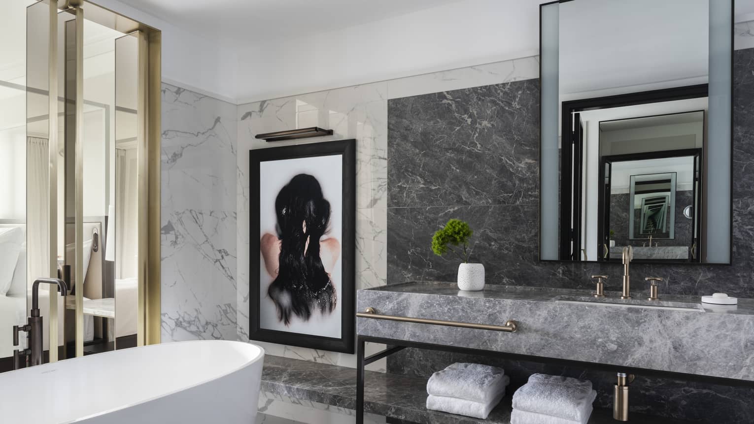 A free-standing tub, grey marble vanity and a framed work of art are set beside glass doors that open to the bedroom