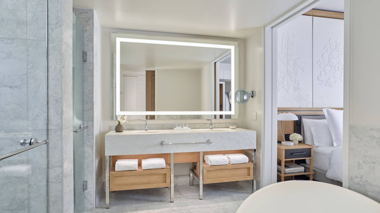 White marble bathroom with light wood shelves, illuminated mirror and freestanding tub