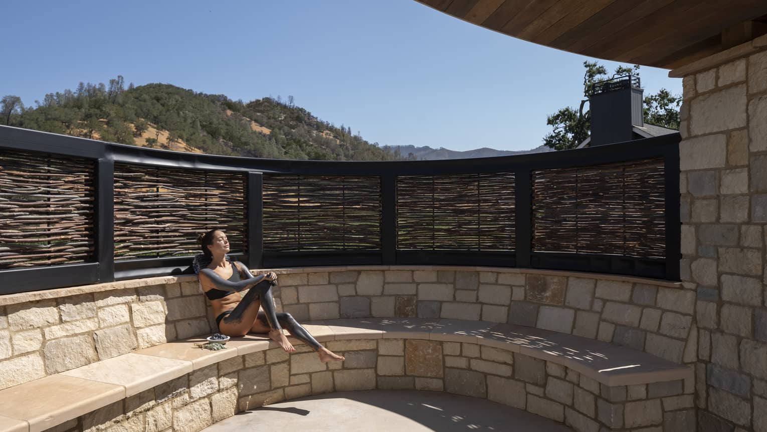Woman slathered in mud sits on stone bench sun tanning during spa treatment