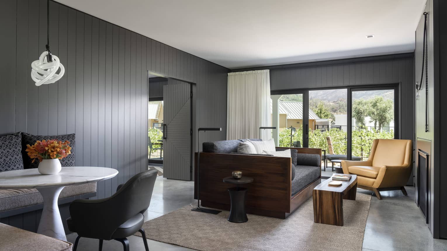 Resort suite living area with a gray sofa, tan armchair, wooden coffee table, small dining area and large windows overlooking greenery