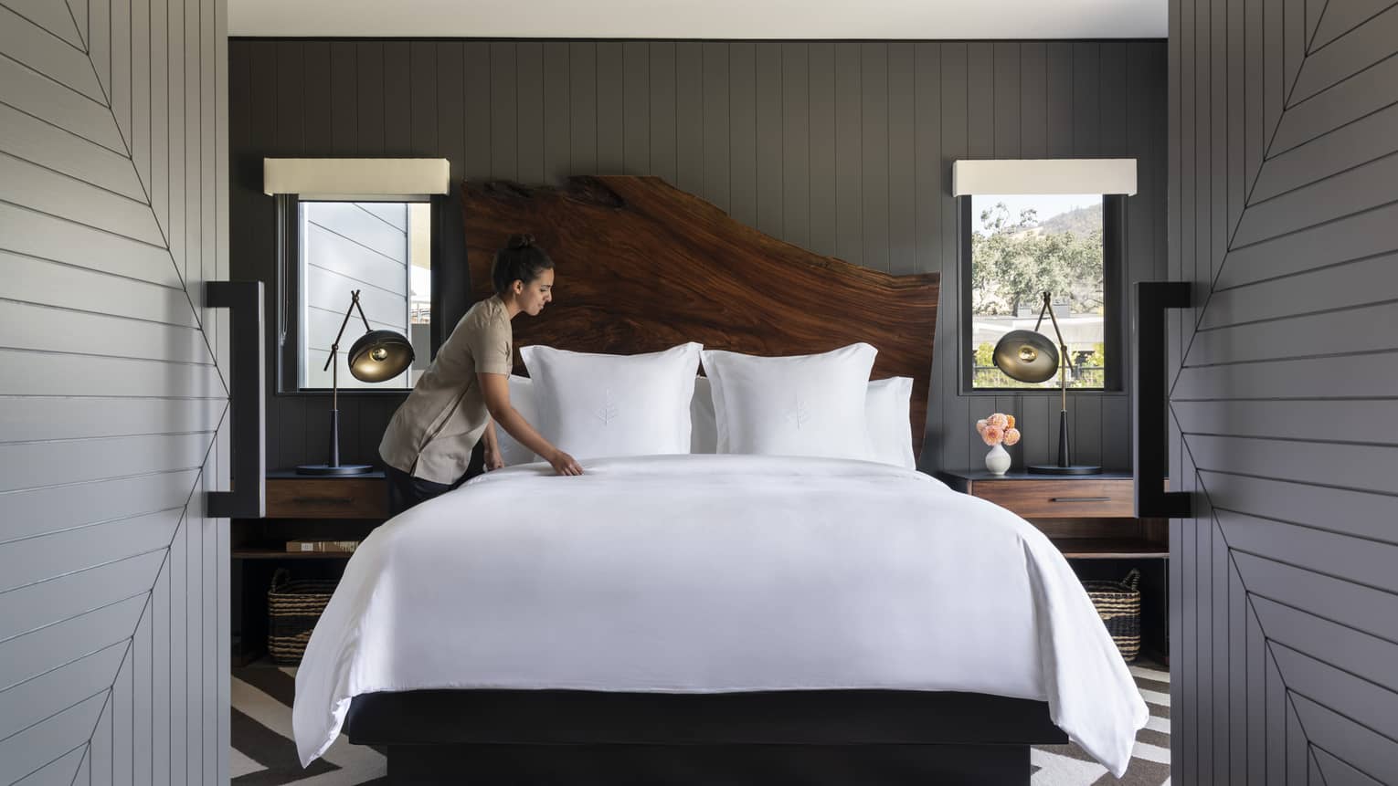 Four Seasons resort staff making bed with white linens in a luxury room featuring a wooden headboard, nightstands with lamps, striped rug and dark paneled walls