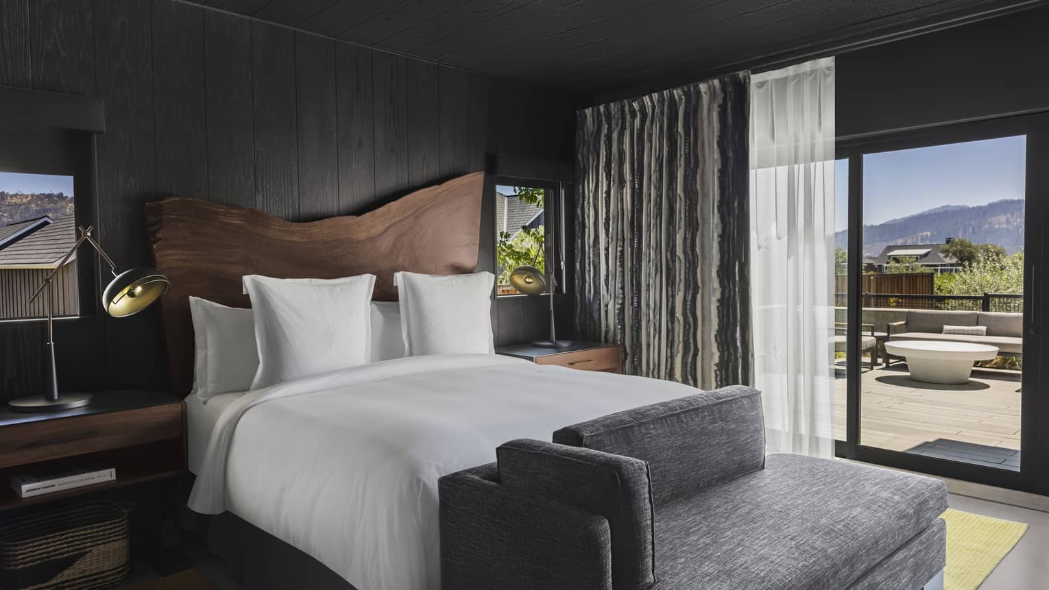 Bedroom with dark wood paneling, king bed, wood headboard, gray chaise, nightstands with lamps and glass doors leading to a private mountain-view terrace 