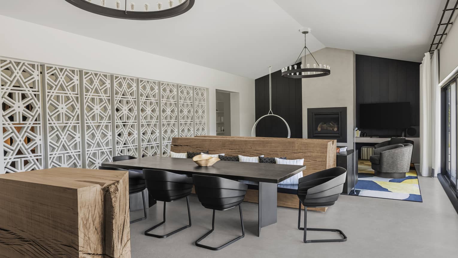 Modern dining area with a black table and chairs, a wooden bench, decorative white lattice wall panels and a view into a living area with a fireplace and TV