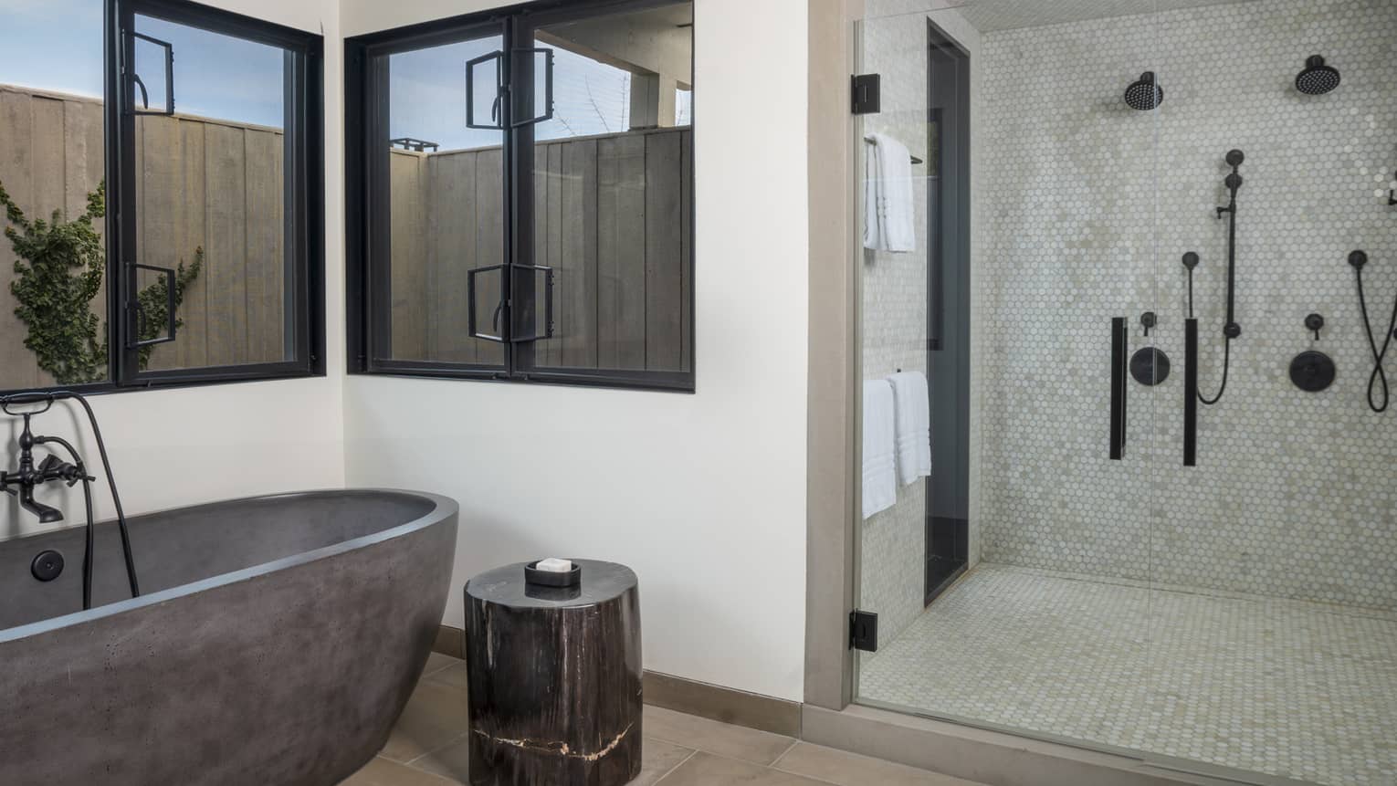 Luxury resort bathroom with a dark freestanding bathtub, wooden side table, glass-enclosed shower with hexagonal tile and windows overlooking an enclosed patio