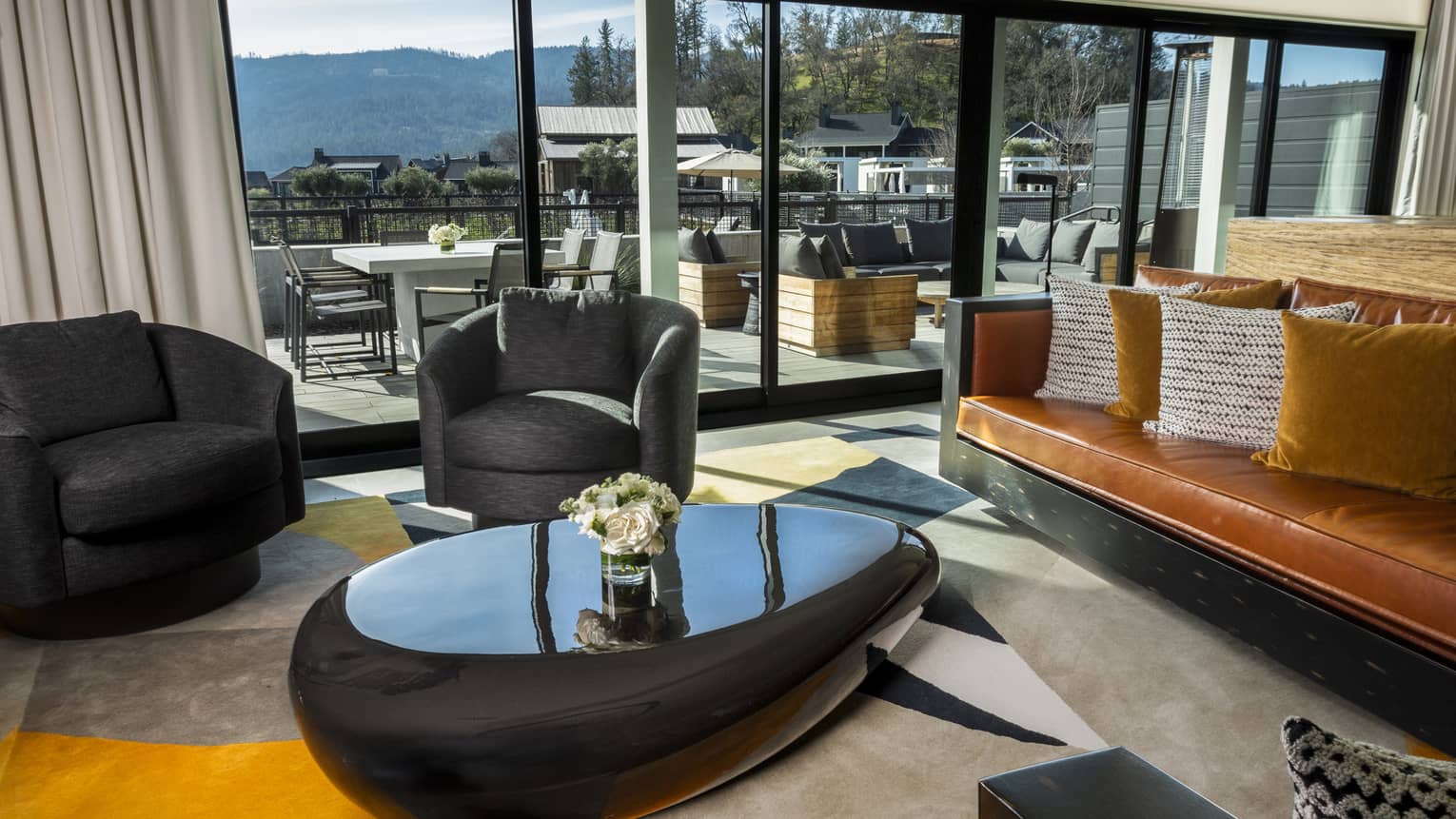 Living room with dark armchairs, caramel leather sofa, patterned cushions, black coffee table and glass doors opening to a spacious terrace with mountain views