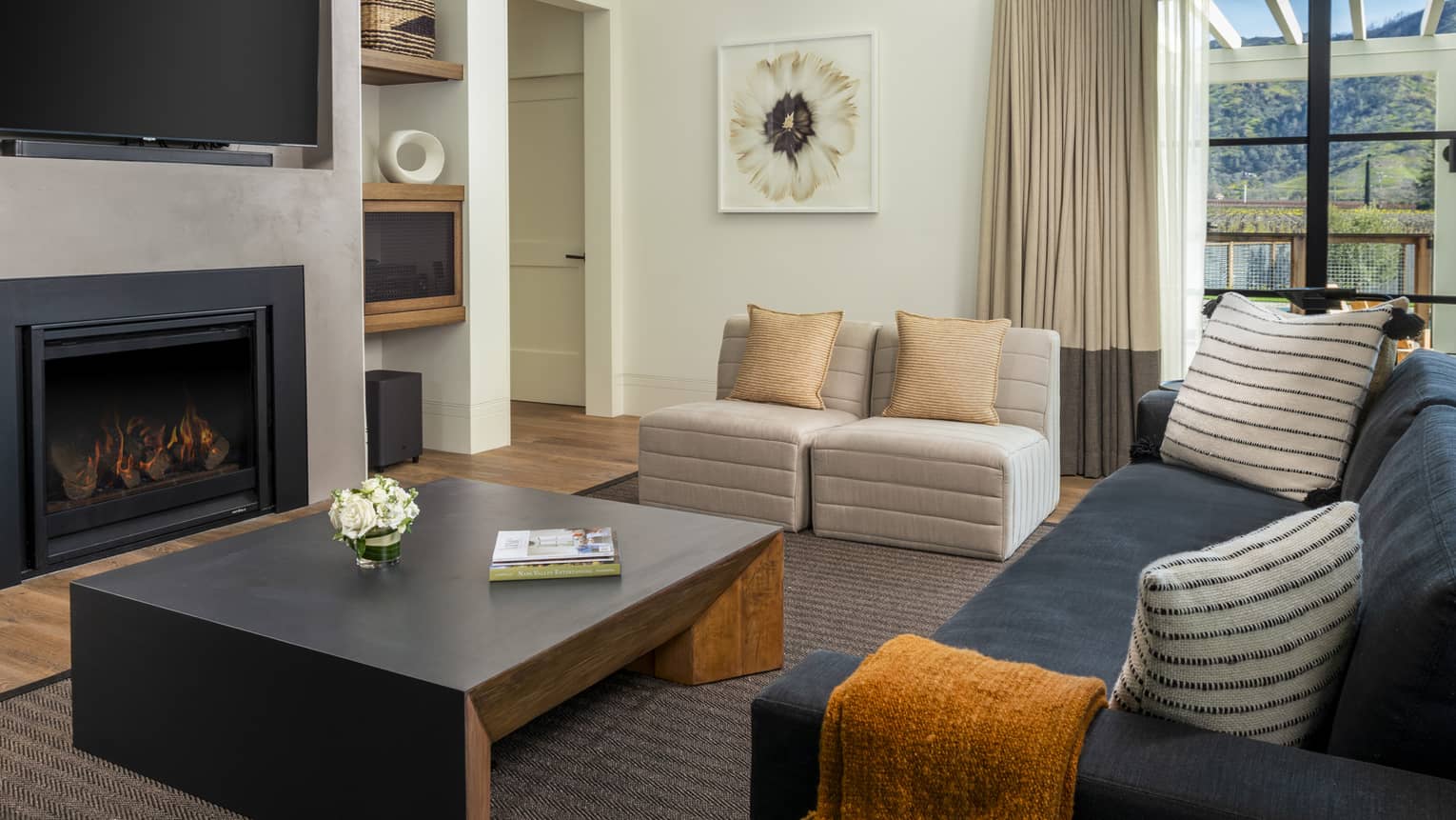 Luxury resort living room with black fireplace, wall-mounted TV, beige armchairs, dark sofa with striped pillows and black coffee table