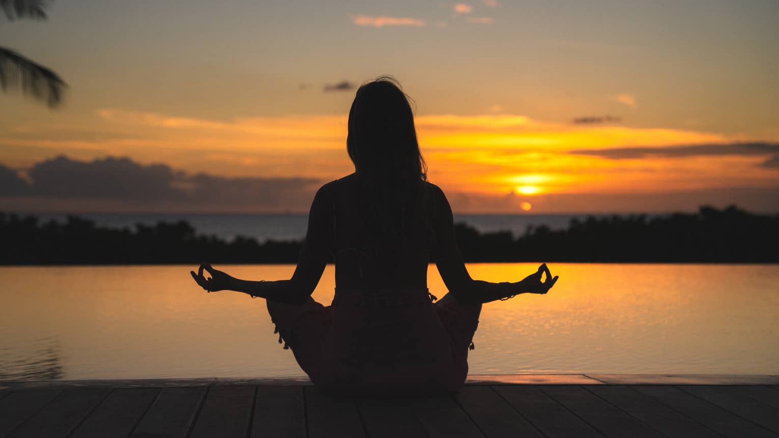 Silhouette of woman in meditation pose in front of calm ocean and orange sunset sky