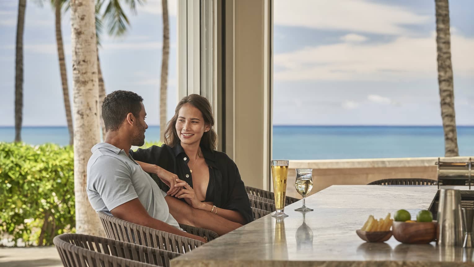 Couple sitting, smiling while seated at open-air bar with ocean in backdrop