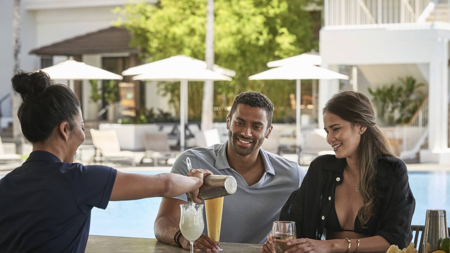 Female bartender pours a tropical drink for man and woman at poolside bar