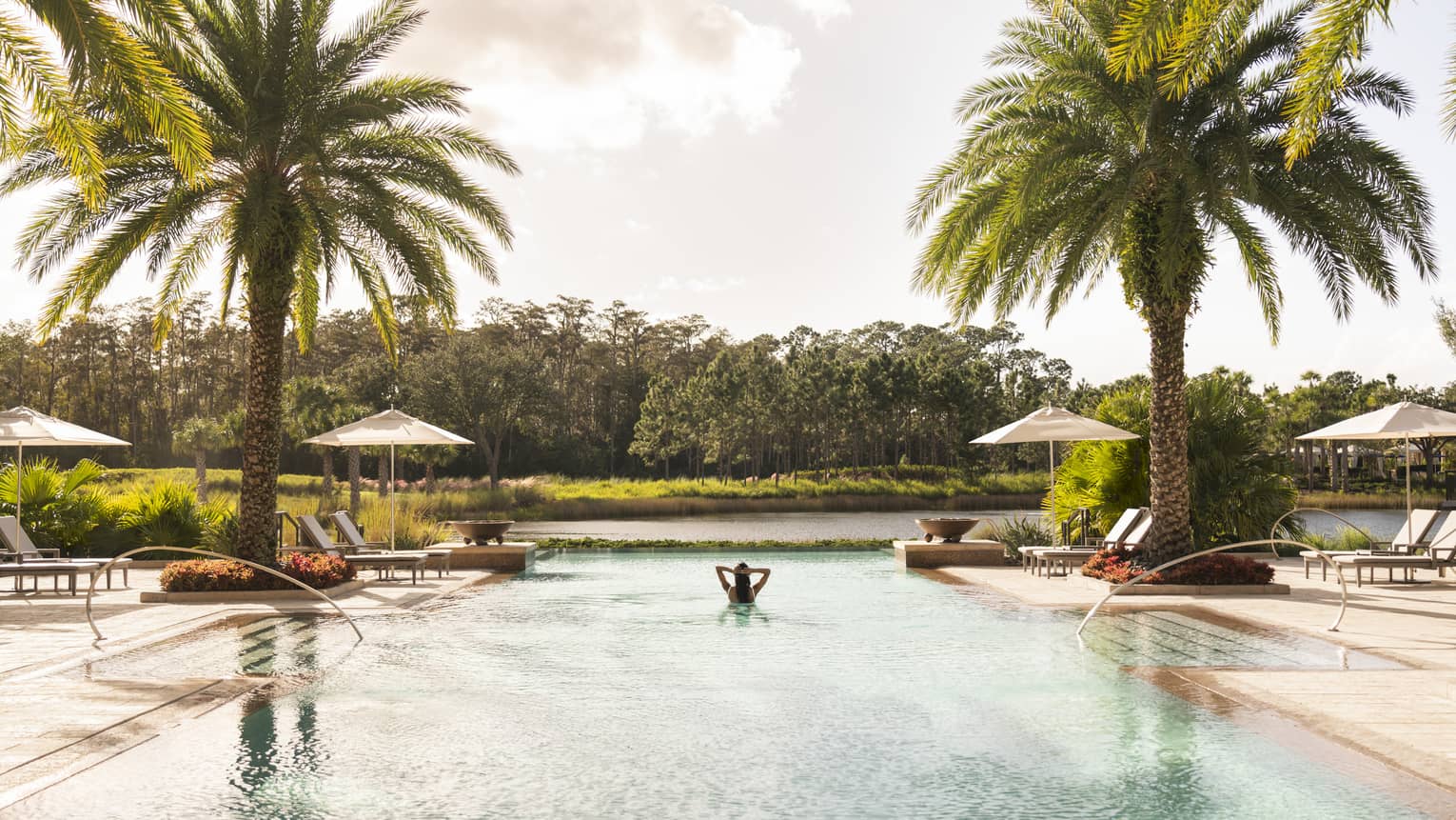 A woman alone in a large pool with lounge chairs and palm trees around it.