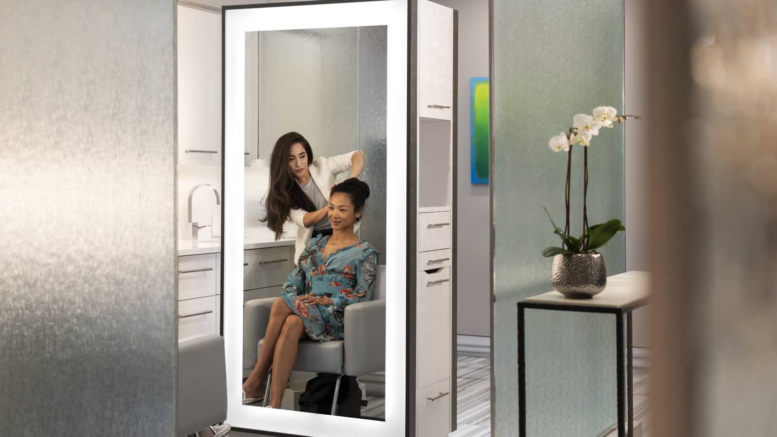 A woman styles another woman's hair; both reflections are seen in an illuminated mirror at salon