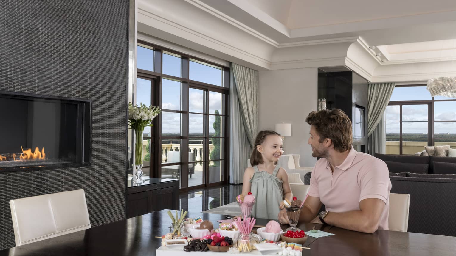Man and young girl sit at dining table in hotel suite, next to fireplace
