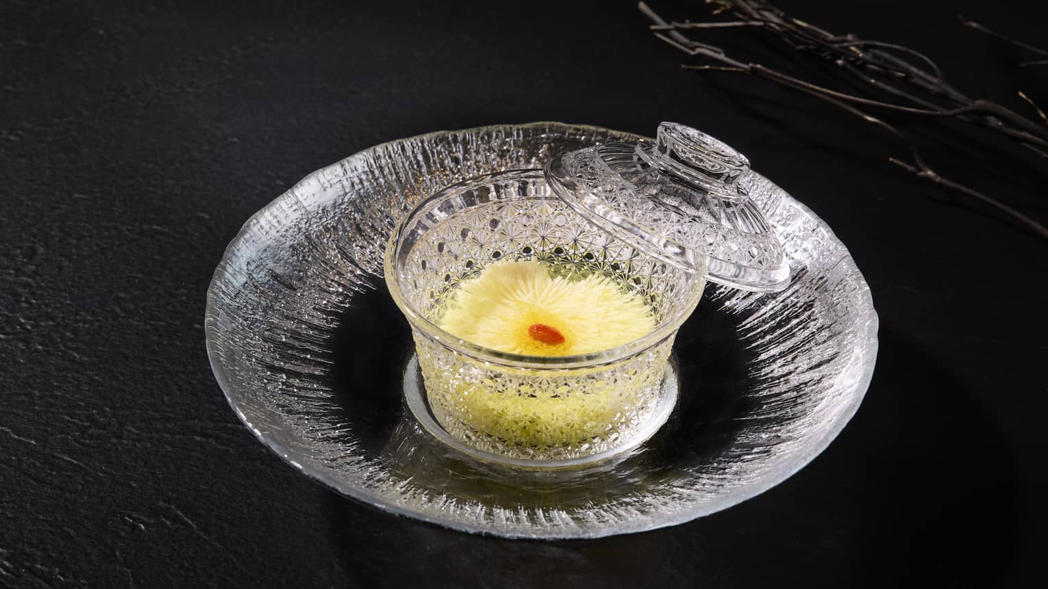 A clear glass plate with a clear glass bowl in the center holds a yellow food item with a small red garnish on top; the bowl?s clear glass lid is propped on the plate against the bowl?s edge.