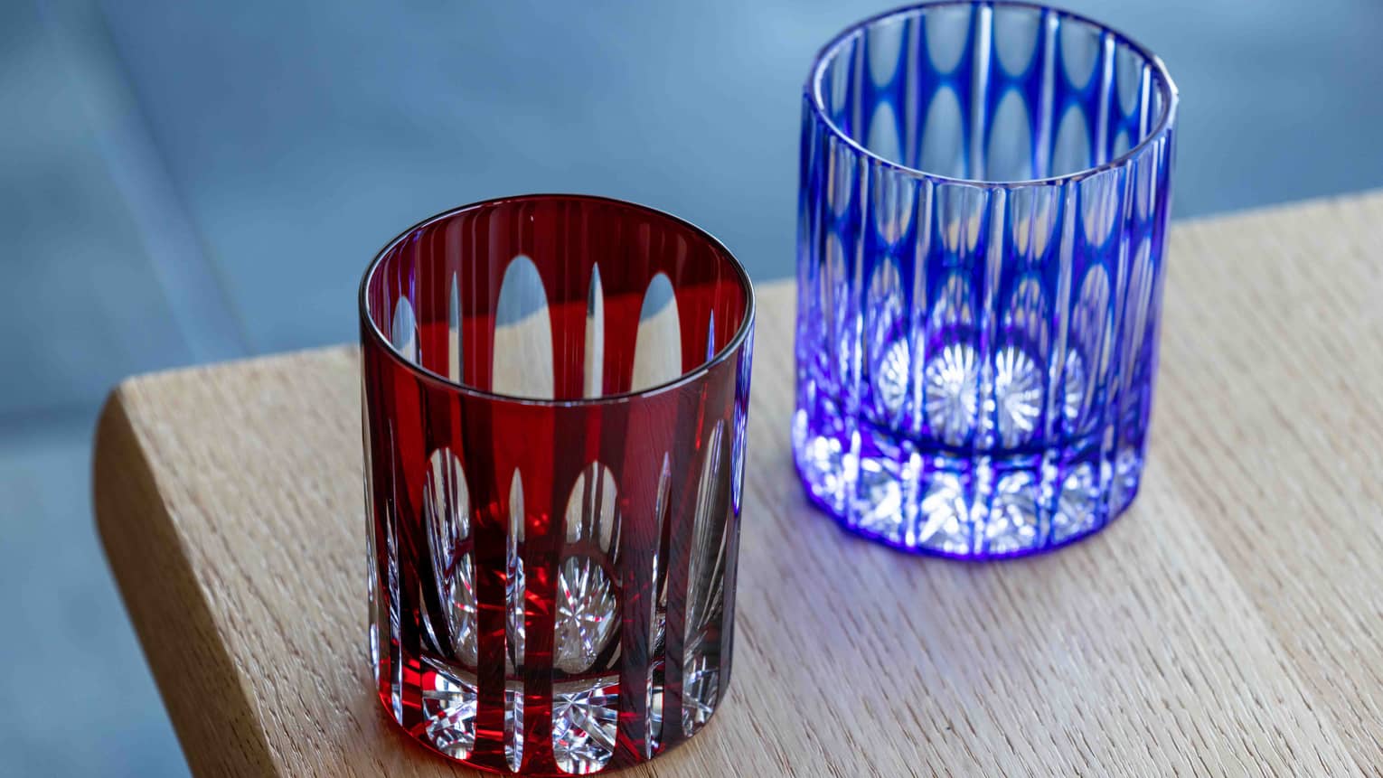 Close-up of two cut-glass tumblers with intricate geometric patterns overlaid by coloured glass, one dark red, one blue.