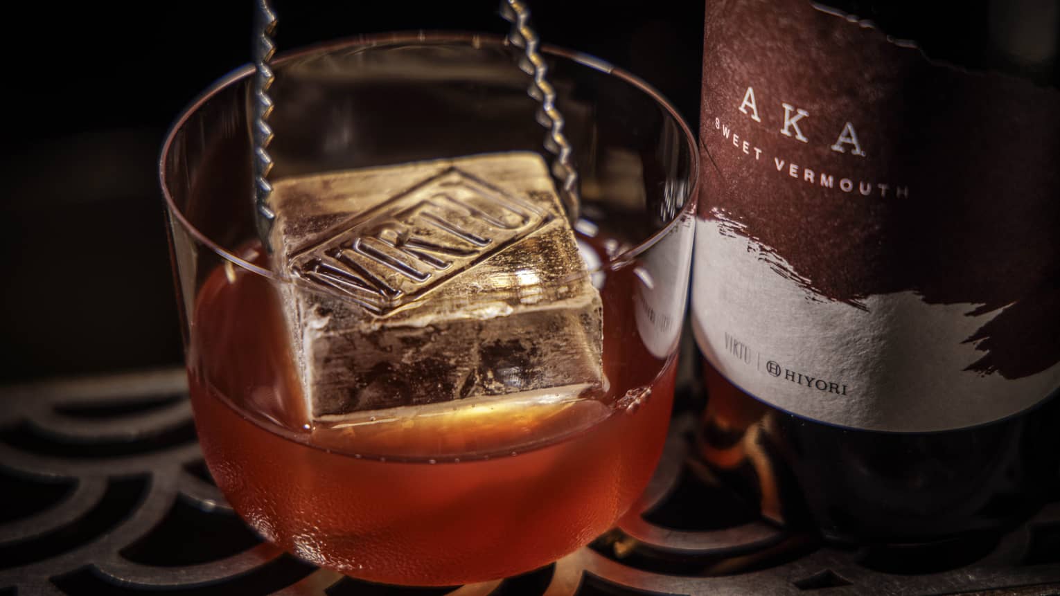 A pair of metal tongs lowers a large square ice cube into a chilled cocktail next to a bottle of Vermouth
