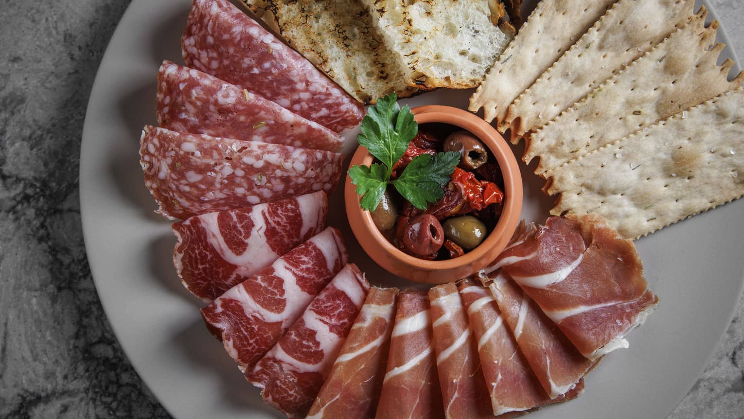 Aerial view of assorted meats, bread and crackers on grey plate with bowl of olives in the centre