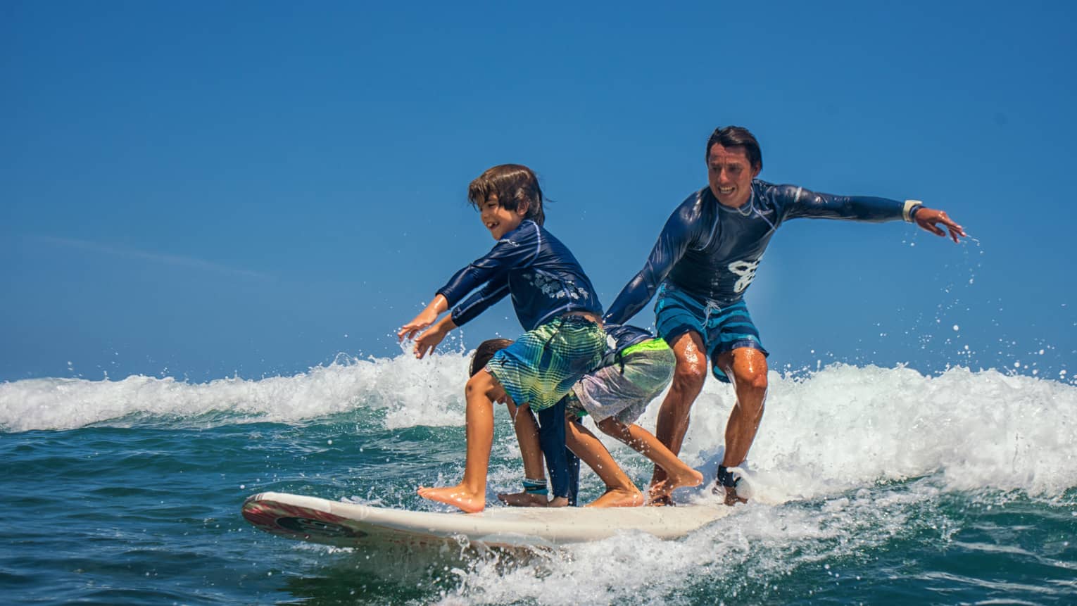 Adult and two children share a surfboard, laughing as they balance on a foamy wave. 