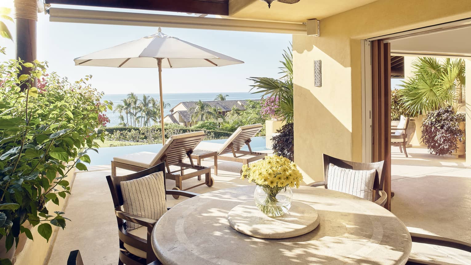 Round dining table with yellow flowers in vase under covered patio, umbrella in sun