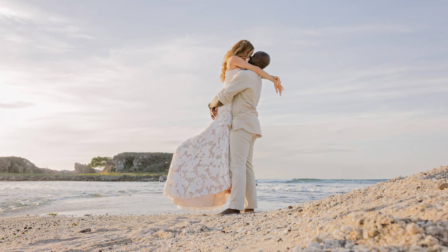 A bride and groom embrace on a beach at sunset