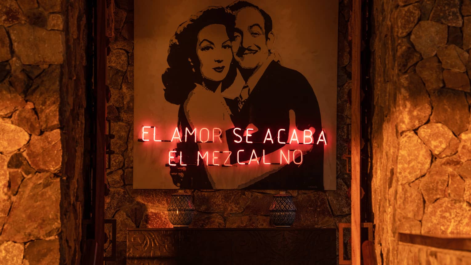 A neon sign with Spanish writing in front of a painting of two people hugging on a stone wall.