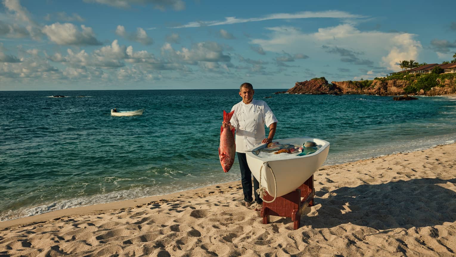 Chef in white chef’s coat, standing on beach beside a small boat, holding a freshly caught fish