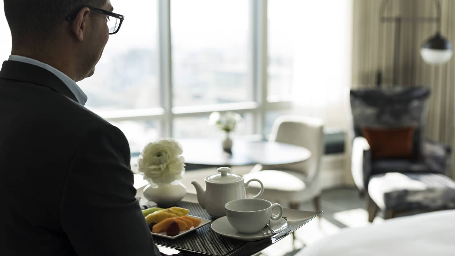 A man carries a tray of tea inside a hotel guest room with window