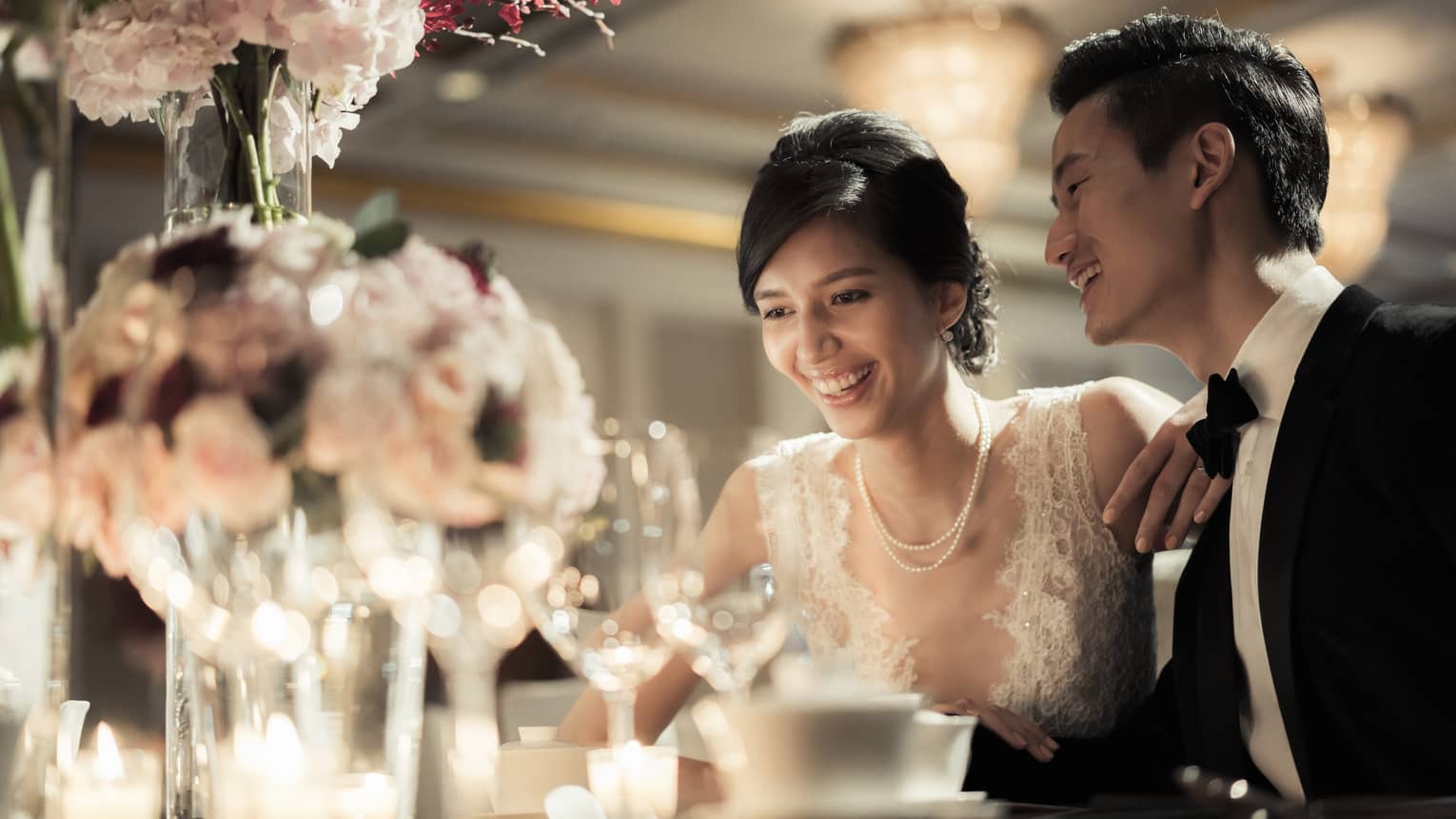 Smiling bride and groom at wedding dining table with glowing candles, pink roses