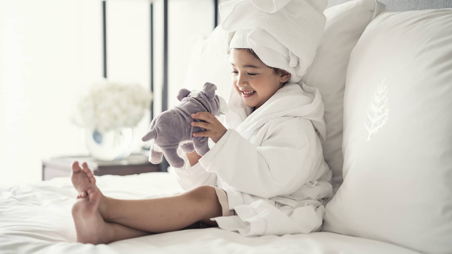 Young girl wearing a white bathrobe with towel wrap plays with plush dog toy on bed