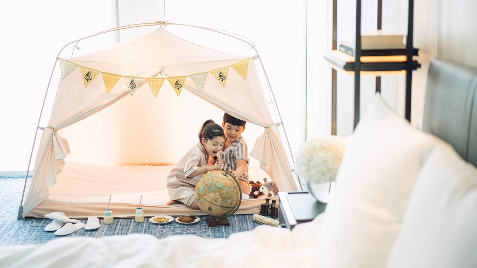 Two children wearing white bathrobes play in white tent in hotel room by bed