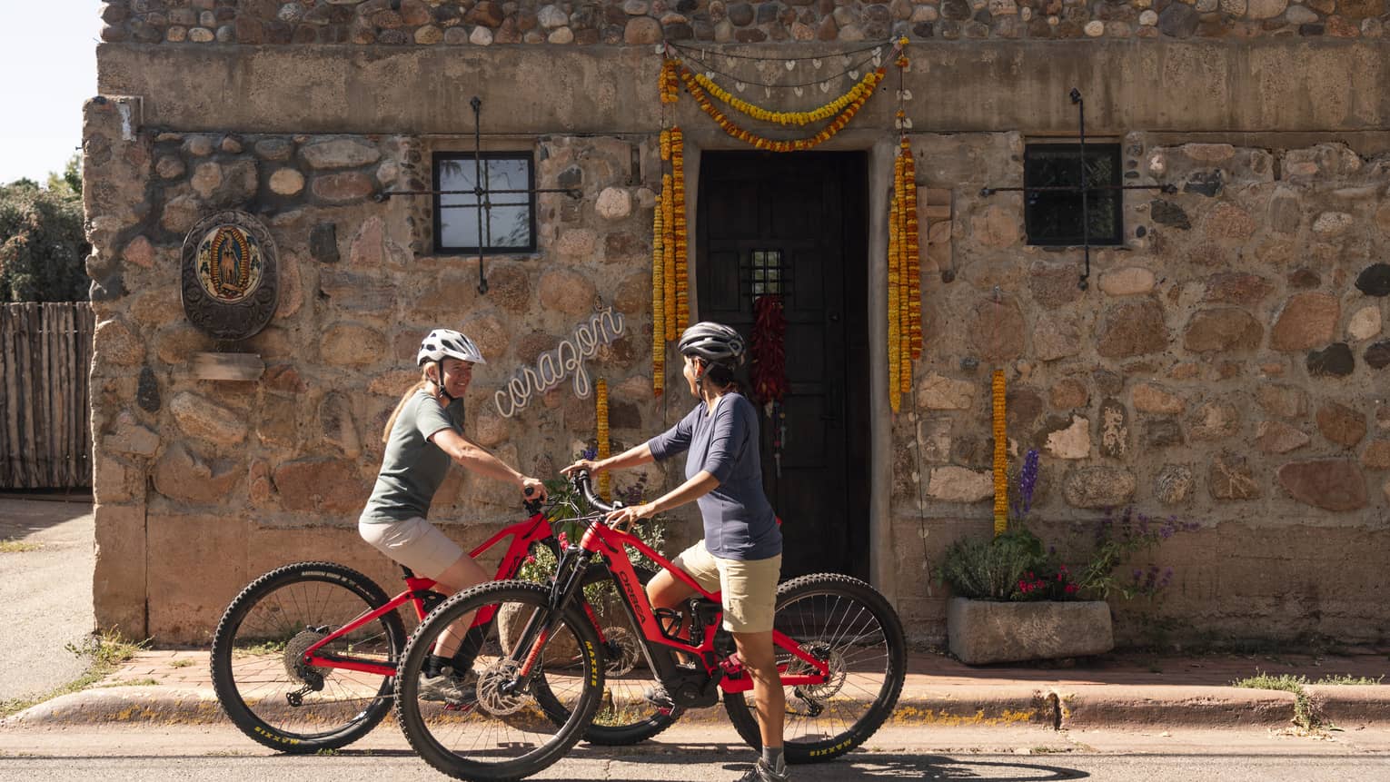 Two women on electric bikes outside of an old building.
