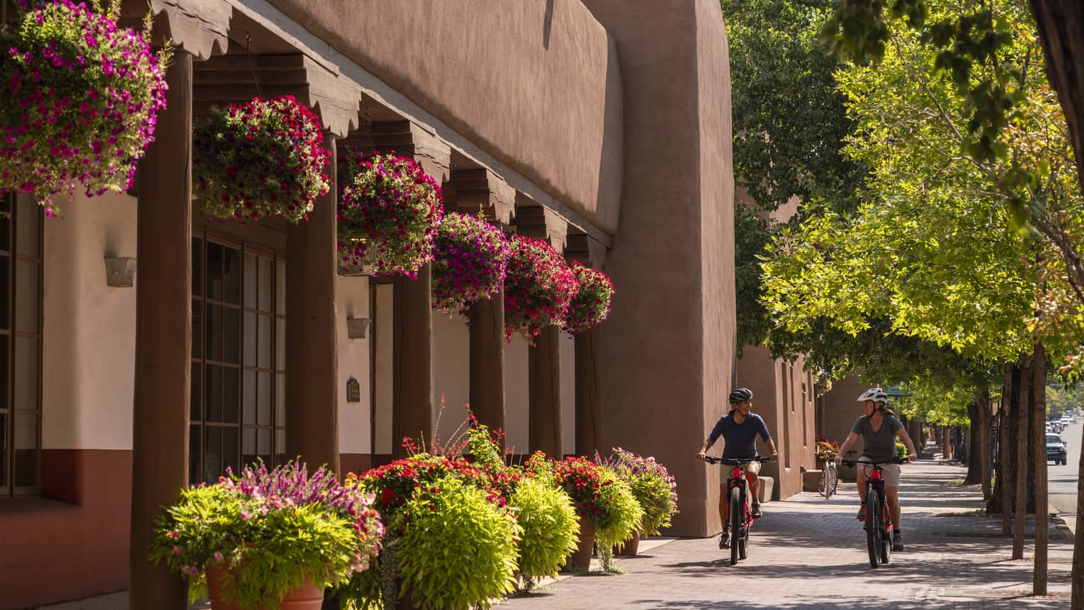 Two people on bikes riding down a small street with a building on one side and small trees on the other.