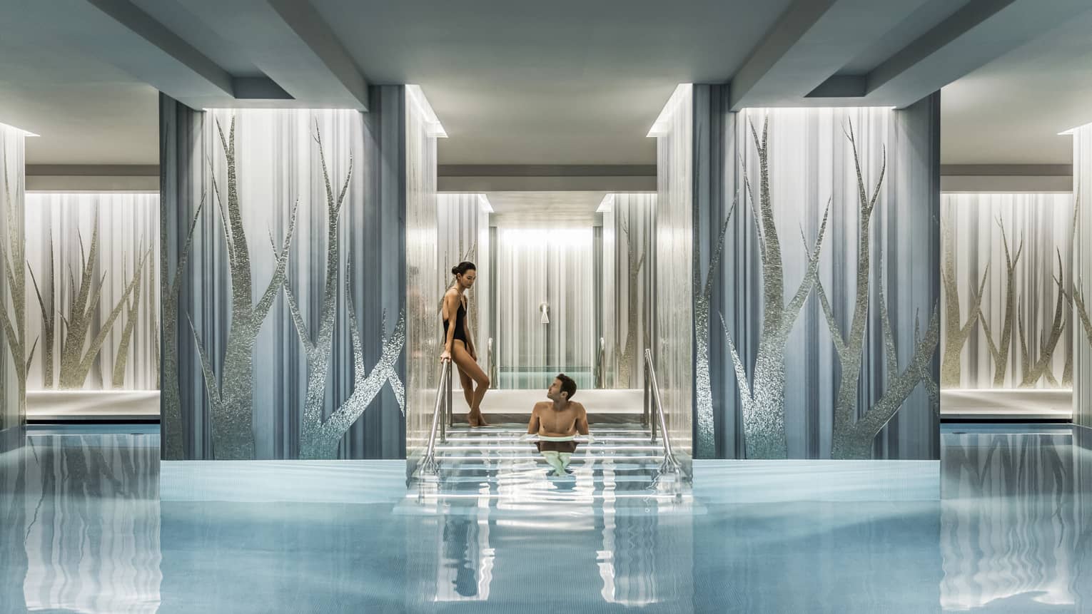A couple enjoys a serene swimming pool, with one person seated on steps between elegant blue and silver columns.