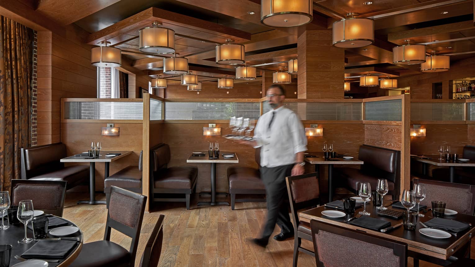 A team member walking with a tray of glasses through dimly lit restaurant with wooden floor and tables.