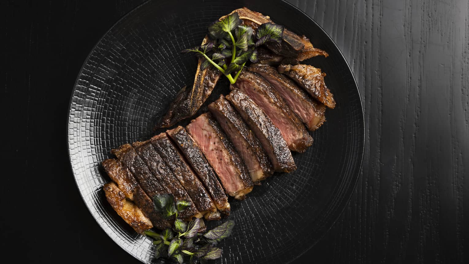 A large steak sliced into thin strips, garnished with leafy greens, served on a black ceramic plate