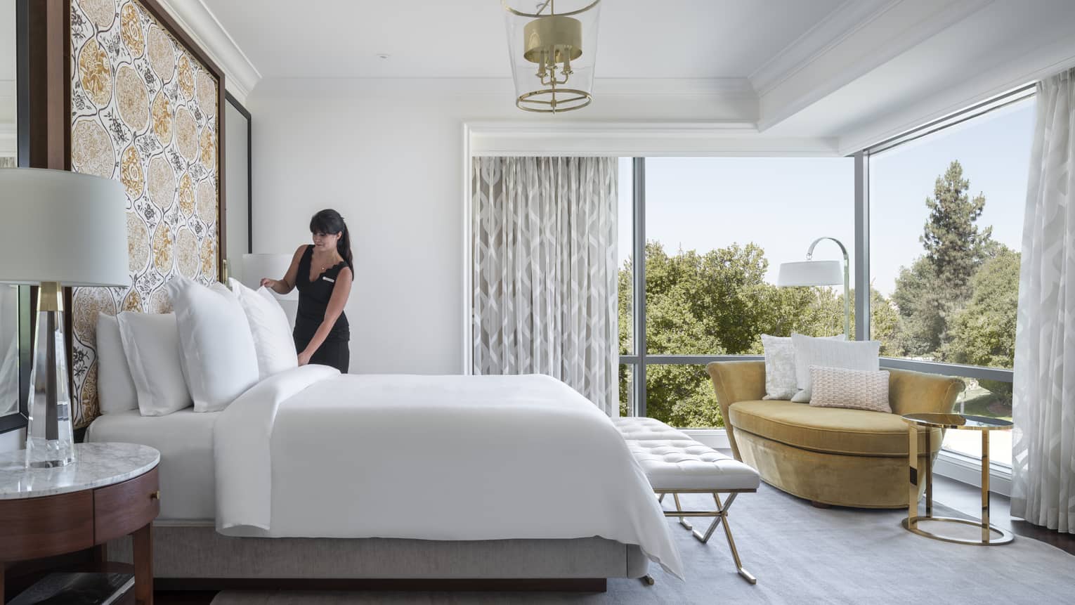 A woman tidying up a bed in a brightly lit hotel room with large windows.
