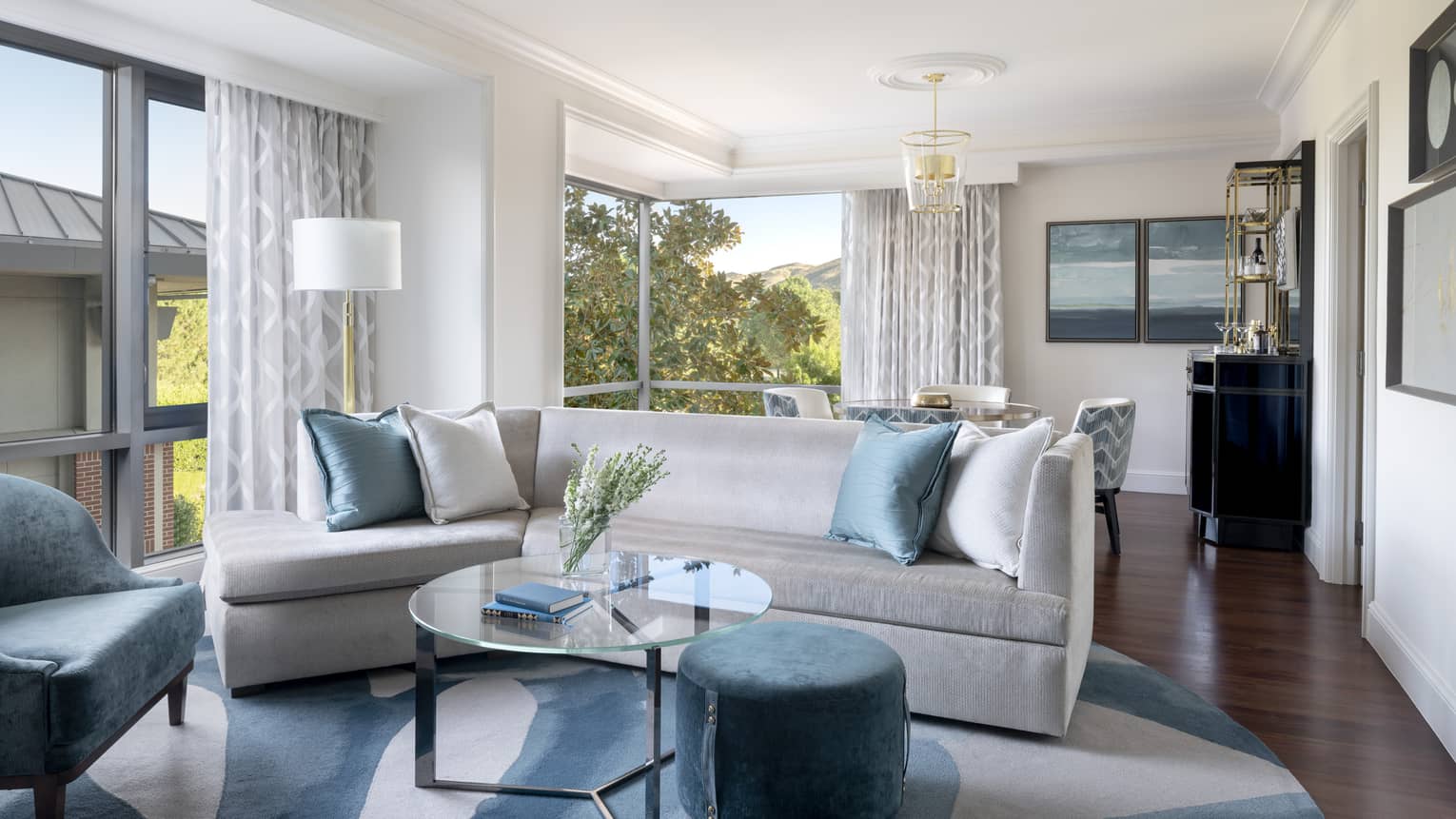 Silver couch with blue pillows, a blue chair, glass coffee table, and a view of trees 