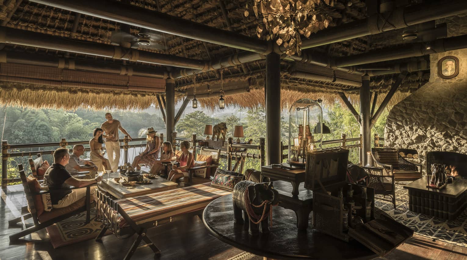 People mingle at open-air bar, beamed thatched roof, lounge seating, trees in the distance