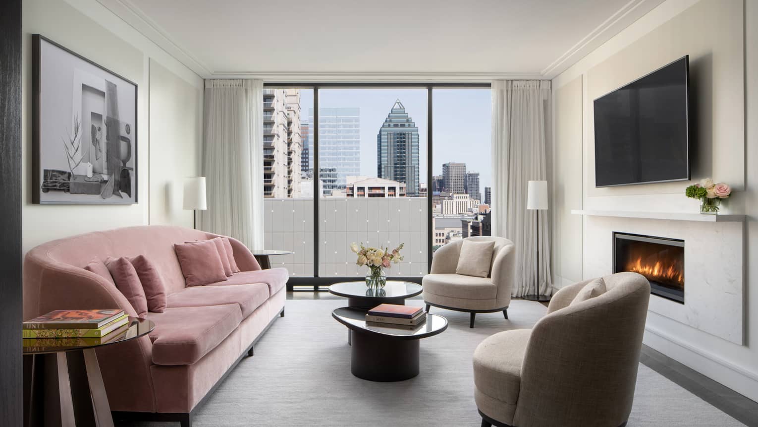 Hotel living room with pink velvet sofa, two arm chairs, fireplace, TV and city view